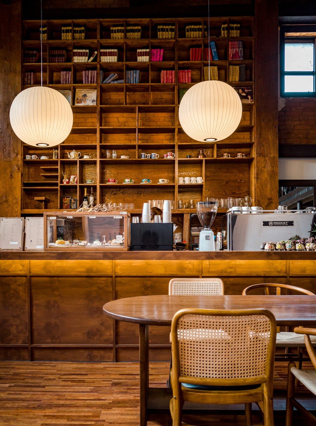The Ember and Oak interior at the corner counter, wooden bookshelves stacked with mugs and books, two white globe pendant lamps over the espresso bar.
